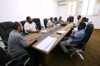 Group of men in business casual sit around a long conference table in a meeting room on a workday (informative scene).