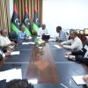 Group of professionals seated around a long conference table discussing; three Libyan flags stand in the background behind them.