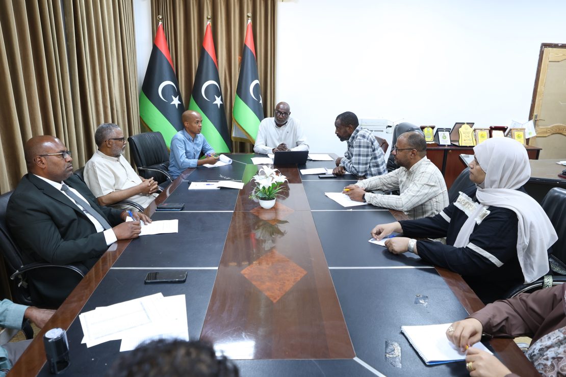 Group of professionals seated around a long conference table discussing; three Libyan flags stand in the background behind them.
