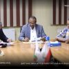 Three men in business attire sit at a conference table reviewing documents, with flags visible in the foreground and an Arabic caption across the bottom.