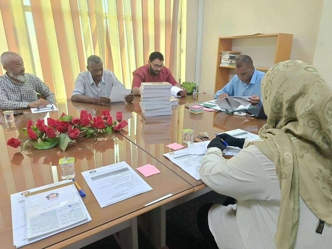 Five adults seated around a glass conference table reviewing documents, with a tall stack of books and pink flowers in the foreground.