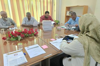 Five adults seated around a glass conference table reviewing documents, with a tall stack of books and pink flowers in the foreground.