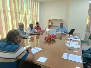 Group of men seated around a glossy conference table, discussing documents under bright, sunlit windows with vertical blinds.