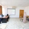 Woman in a hijab gives a presentation to three men seated at a desk in a classroom with a projector screen behind them and a whiteboard on the wall.