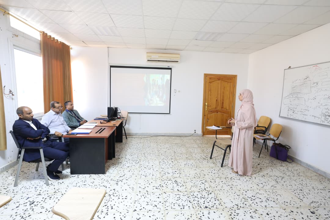 Woman in a hijab gives a presentation to three men seated at a desk in a classroom with a projector screen behind them and a whiteboard on the wall.