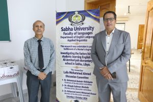 IMG-20261004-WA0080 - جامعة سبها | Sebha University Two men in business attire pose beside a tall Sabha University banner announcing a language department event.