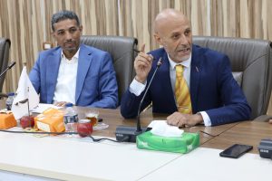 Two men in blue suits sit at a conference table; the man on the right speaks into a microphone while gesturing with a pen.",