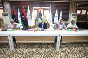 Panel of five men seated at a conference table with multiple national and organizational flags behind them.