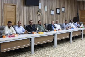 IMG-20261008-WA0070 - جامعة سبها | Sebha University Nine men sit along a long conference table in a wood-paneled meeting room, with water bottles and snacks on the table.