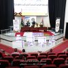 Auditorium stage with two women speakers behind a podium, banners and Libyan flag, awards on the table, and gift boxes arranged on the red-carpeted floor behind an attentive audience.