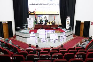 Auditorium stage with two women speakers behind a podium, banners and Libyan flag, awards on the table, and gift boxes arranged on the red-carpeted floor behind an attentive audience.