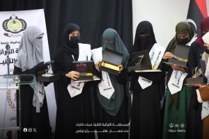 Women in hijab in graduation gowns receive certificates on stage at a university ceremony, with a banner and logo in the background.