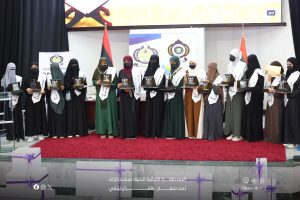 Group of women in hijabs and abayas on a stage, each holding an award plaque during a ceremony, with banners and flags in the background.