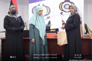 Three women in dark abayas stand on a stage during an award ceremony; one hands a certificate to another, with gift bags nearby and banners in the background.
