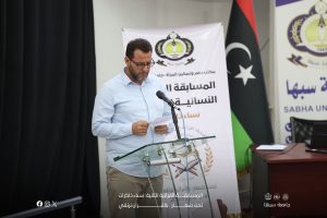 Man in a light blue shirt speaks at a glass podium with a microphone, Arabic banners behind him.