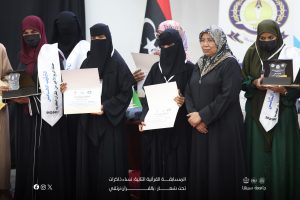 Group of women in hijabs wearing black abayas receiving certificates and glass trophies at an awards ceremony with a logo backdrop.