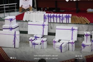 White gift boxes and paper bags wrapped with purple ribbons arranged on marble steps at a stage, likely for a Quran competition event (Arabic banners visible).