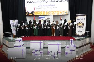 Group of female graduates in hijabs on a stage, receiving certificates; banners and a large screen behind them, with wrapped gift boxes in the foreground.