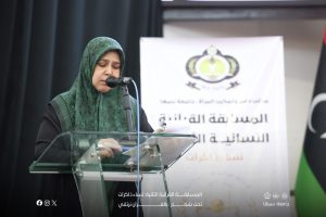 Woman in a green hijab speaks into a microphone at a glass podium, with an event banner behind her.