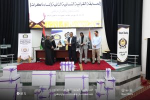 حفل توزيع جوائز لمسابقة قرآنية على منصة أمام جمهور، مع متسابقين ومتلقّين الجوائز بجانب علم شعار الجامعة