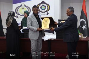 Man in a light gray suit receives a decorative plaque from a man in a dark suit at an awards ceremony, with onlookers in the background and flags on the side