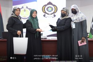 Four women in black abayas at a formal ceremony; two exchange a certificate while others stand nearby with banners and logos in the background.