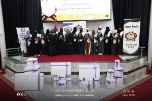 Group of women in graduation robes holding plaques on a stage during an award ceremony, with banners, flags, and wrapped gifts in the foreground on a red carpeted platform.