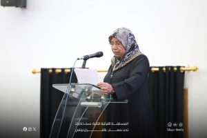 Woman in a patterned hijab and glasses speaks into a microphone at a glass podium, reading from a paper.