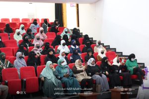 Group of women wearing hijabs and face masks seated in red auditorium chairs at a conference or presentation.