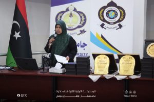 Woman in a green hijab speaks into a microphone at a podium during an awards ceremony, with university banners and the Libyan flag in the background.