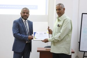 Two men in an office exchange a certificate; one wears a blue suit, the other a light green shirt/coat.
