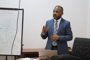 Formal presenter wearing a blue suit and striped tie speaks while gesturing near a whiteboard with diagrams, laptop open on a table behind him