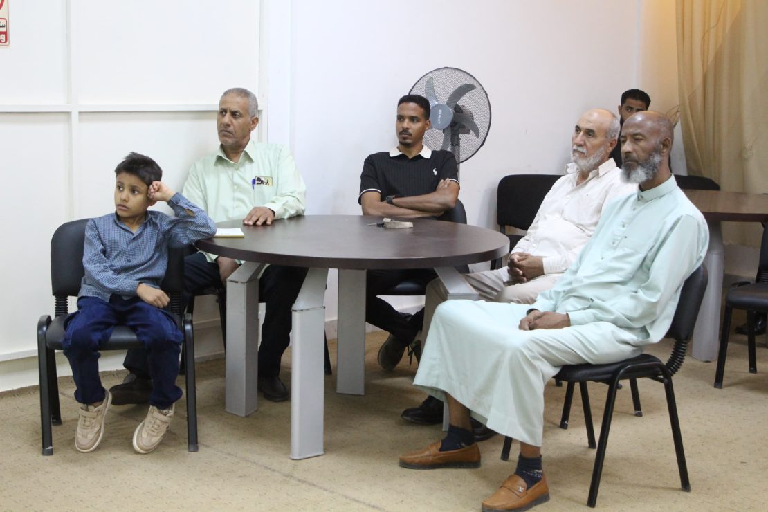 Six people, including a boy in a wheelchair, sit around a round table in a meeting room framing a casual group gathering.