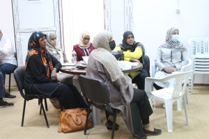 Group of women wearing hijabs seated around a round table in a bright meeting room, some with masks and papers on the table.