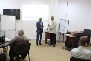 Two men shake hands as one receives a certificate during a presentation in a conference room with attendees watching.