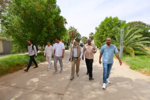 IMG-20261011-WA0003 - جامعة سبها | Sebha University Group of men walking together down a sunny path flanked by green trees and palms, engaged in conversation. Seven adults in light-colored shirts and trousers.