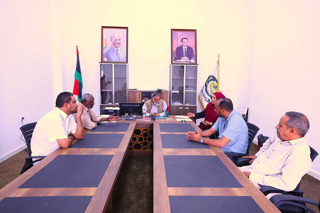 People sit around a large conference table in a meeting room, gesturing and talking; flags and framed portraits are visible in the background.
