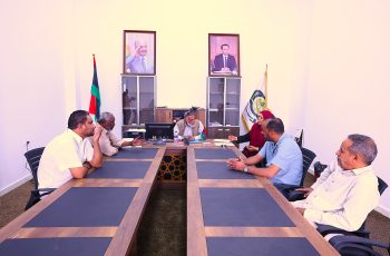 People sit around a large conference table in a meeting room, gesturing and talking; flags and framed portraits are visible in the background.