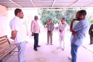 IMG-20261011-WA0006 - جامعة سبها | Sebha University Six men stand in a semicircle under a metal roof, engaged in a discussion outdoors on a sunny day.