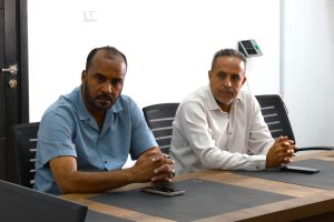 Two men sit at a conference table in a meeting room, looking serious and focused.