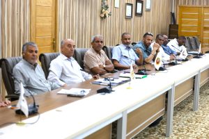 IMG-20261011-WA0011 - جامعة سبها | Sebha University Group of men seated along a long conference table in a wood-paneled room, with microphones and small flags on the table.