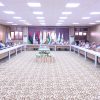 Formal meeting in a conference room with delegates seated around a U-shaped table and multiple flags displayed at the back of the room.]','