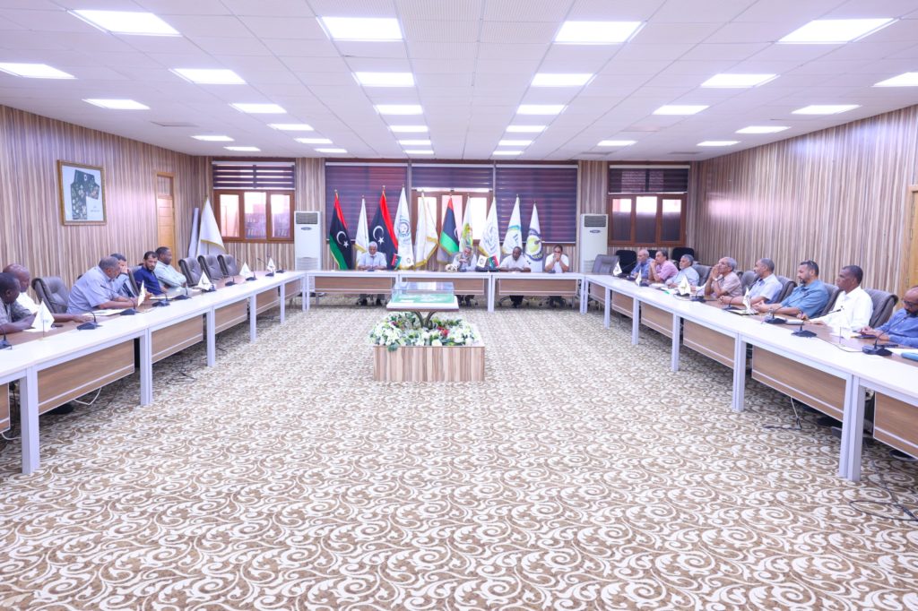 Formal meeting in a conference room with delegates seated around a U-shaped table and multiple flags displayed at the back of the room.]','
