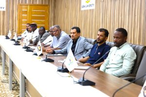 IMG-20261011-WA0014 - جامعة سبها | Sebha University Group of men seated at a long conference table with microphones, flags, and a wooden paneled wall in a formal meeting room.