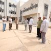 Six men in casual attire stand in a semicircle in a dusty courtyard outside a beige building with many windows and wall-mounted AC units.