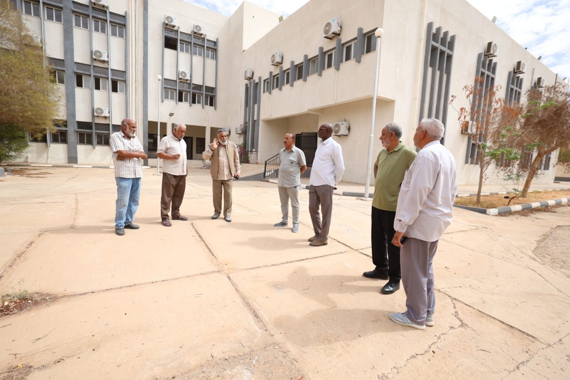 Six men in casual attire stand in a semicircle in a dusty courtyard outside a beige building with many windows and wall-mounted AC units.