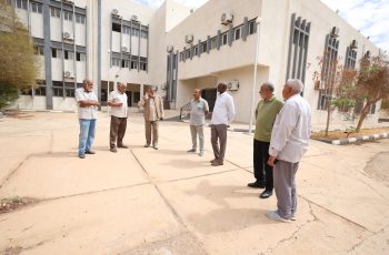 Six men in casual attire stand in a semicircle in a dusty courtyard outside a beige building with many windows and wall-mounted AC units.