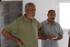 Two men stand side by side indoors; one in a green polo points or rests a hand on his hip while the other in gray looks on and writes notes on paper.