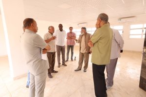 Group of men standing in a circle, conversing in a bright indoor room with tiled floor and windows on the right.