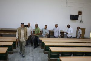Group of men walking through a classroom with long wooden desks and green chairs far wall light gray.